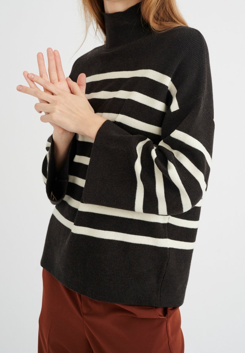 Suéter negro de punto con rayas horizontales blancas, cuello alto y mangas acampanadas, combinado con pantalones color óxido. Textura suave, diseño informal.