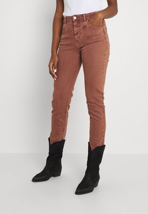 Slim-fit braune Jeans, getragen mit schwarzen Cowboy-Stiefeln bis zur Mitte der Wade, Modell steht mit einer Hand in der Tasche und weißem, eingestecktem Oberteil.