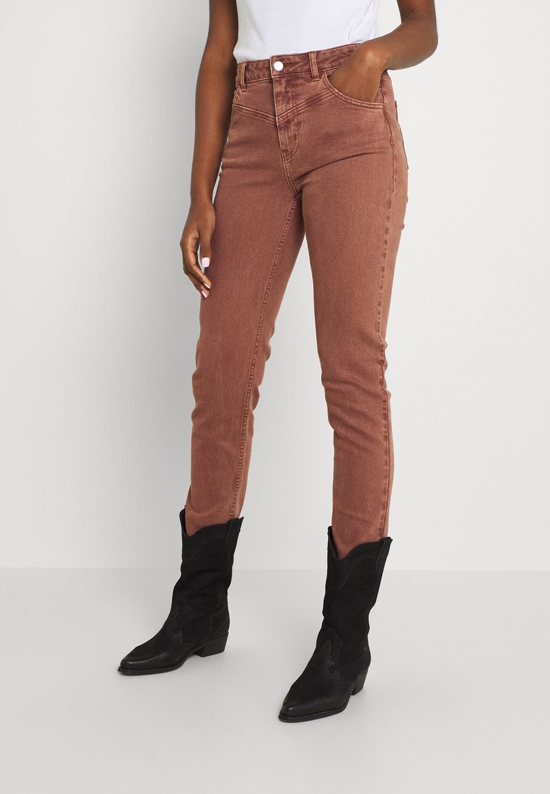 Jean marron slim porté avec des bottes de cowboy noires mi-mollet, modèle debout avec une main dans la poche et un top blanc rentré.
