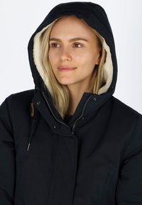 Giacca nera isolante con cappuccio, caratterizzata da un morbido rivestimento crema, chiusura con zip e sottili dettagli in legno sulla chiusura.