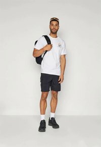 Λευκό μπλουζάκι με πολύχρωμο λογότυπο, μαύρες cargo βερμούδες και μαύρα αθλητικά παπούτσια. Το άτομο κουβαλάει μια μαύρη πλάτη και φοράει γυαλιά ηλίου.