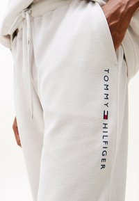 Weiße Baumwoll-Jogginghosen mit glatter Textur. Seitentasche sichtbar. "TOMMY HILFIGER"-Logo vertikal in Blau auf dem linken Bein aufgedruckt.
