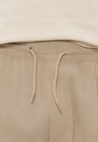 Μπεζ λινό παντελόνι με ελαστική μέση και ταιριαστά κορδόνια, φορεμένο με ανοιχτό μπεζ τοπ.