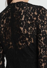 Musta pitsimekko lillemuster med lange ærmer og høj halsudskæring. Har en kompleks tekstur med gennemsigtige og faste sektioner.