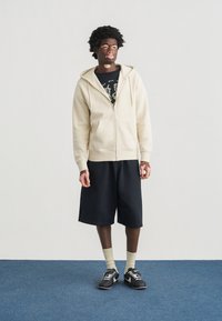 Μπεζ φούτερ με φερμουάρ και μπροστινές τσέπες, μαύρο μπλουζάκι με στάμπα από κάτω, μαύρη βερμούδα, κιτρινοπράσινες κάλτσες και μαύρα αθλητικά παπούτσια.