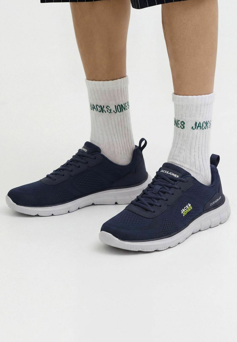 Jambes portant des chaussettes blanches Jack & Jones et des baskets bleu marine Jack & Jones avec des semelles blanches sur fond blanc.