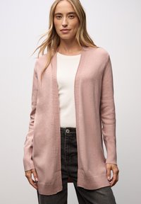 Ljusrosa cardigan gjord av mjukt material, med ribbad öppen front, långa ärmar och en avslappnad passform över en vit topp och grå byxor.