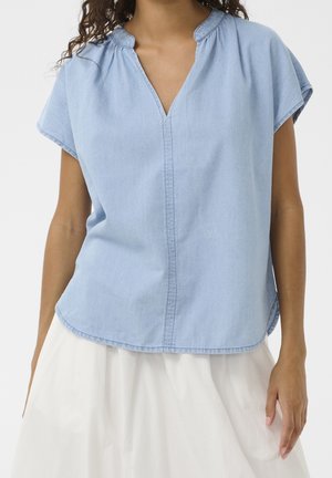 Woman wearing a light blue short-sleeve V-neck top and a white skirt against a plain background.