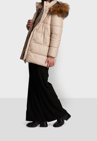 Beige puffermantel med en huva klädd i päls, med dragkedja, sidofickor och en något överdimensionerad passform. Bärs med svarta byxor och stövlar.