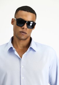 Czarne kwadratowe okulary przeciwsłoneczne z ciemnymi soczewkami, noszone z jasnoniebieską koszulą z kołnierzykiem. Tkanina wygląda gładko z subtelnym połyskiem.