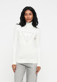 Donna con capelli neri che indossa un maglione bianco a collo alto con un logo triangolare "GUESS" e pantaloni grigio chiaro, che si trova di fronte a uno sfondo bianco.