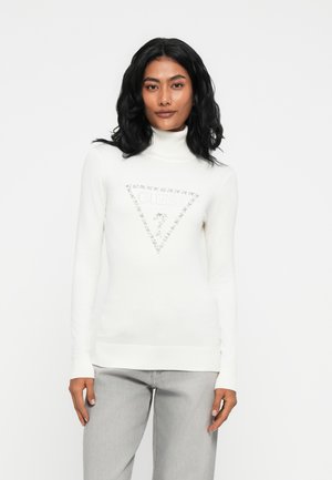 Donna con capelli neri che indossa un maglione bianco a collo alto con un logo triangolare "GUESS" e pantaloni grigio chiaro, che si trova di fronte a uno sfondo bianco.