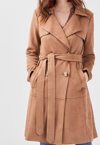 Tan faux suede trench coat with a double-breasted front, belted waist, large pockets, and notch lapels. Smooth texture, knee-length design.