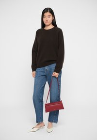 Brun strikket sweater, blå lige ben jeans, hvide spidse flade sko og en rød crossbody taske med et slankt design. Hvid baggrund.