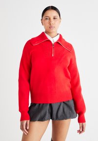 Maglione rosso con zip, texture a coste, colletto oversize e maniche lunghe. Abbinato a pantaloni corti a righe. Sfondo semplice.
