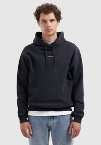 Man met krullend haar draagt een marineblauwe hoodie met de tekst "PURE PATH", lichte spijkerbroek en een wit shirt, staand tegen een effen lichtgrijze achtergrond.