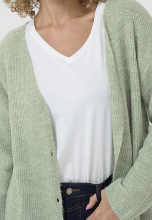 Vrouw draagt een lichtgroen gebreid vest over een wit T-shirt met V-hals en donkerblauwe spijkerbroek, hand in de zak.