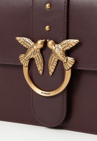 Μπορντό δερμάτινη τσάντα χειρός με κυκλική χρυσή μεταλλική λεπτομέρεια που απεικονίζει δύο λεπτομερείς, ανυψωμένους πουλιά. Περιλαμβάνει χρυσές στρας μπροστά.