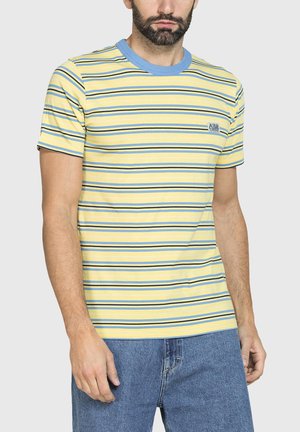 Uomo che indossa una t-shirt gialla a maniche corte con righe orizzontali blu e collo blu, abbinata a jeans di denim blu.