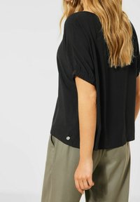 Top corto nero a maniche corte con dettagli arricciati sulle spalle e accenti in metallo argentato; abbinato a pantaloni verde oliva. Tessuto morbido.