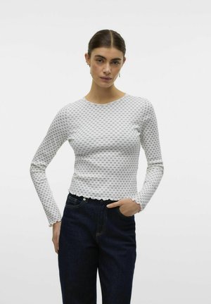 Langærmet tætsiddende top i hvid med et lille gråt mønster, rund halsudskæring og bølget kant, parret med mørke jeans.