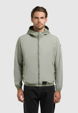 Bărbat care poartă o jachetă cu glugă și fermoar de culoare gri deschis cu manșete elastice și pantaloni negri, stând în fața unui fundal gri simplu.