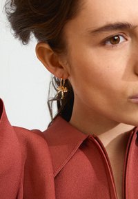 Goldene Creolen mit Schleifenmotiv, getragen zu einem strukturierten, rostfarbenen Oberteil mit Kragen und Reißverschluss.