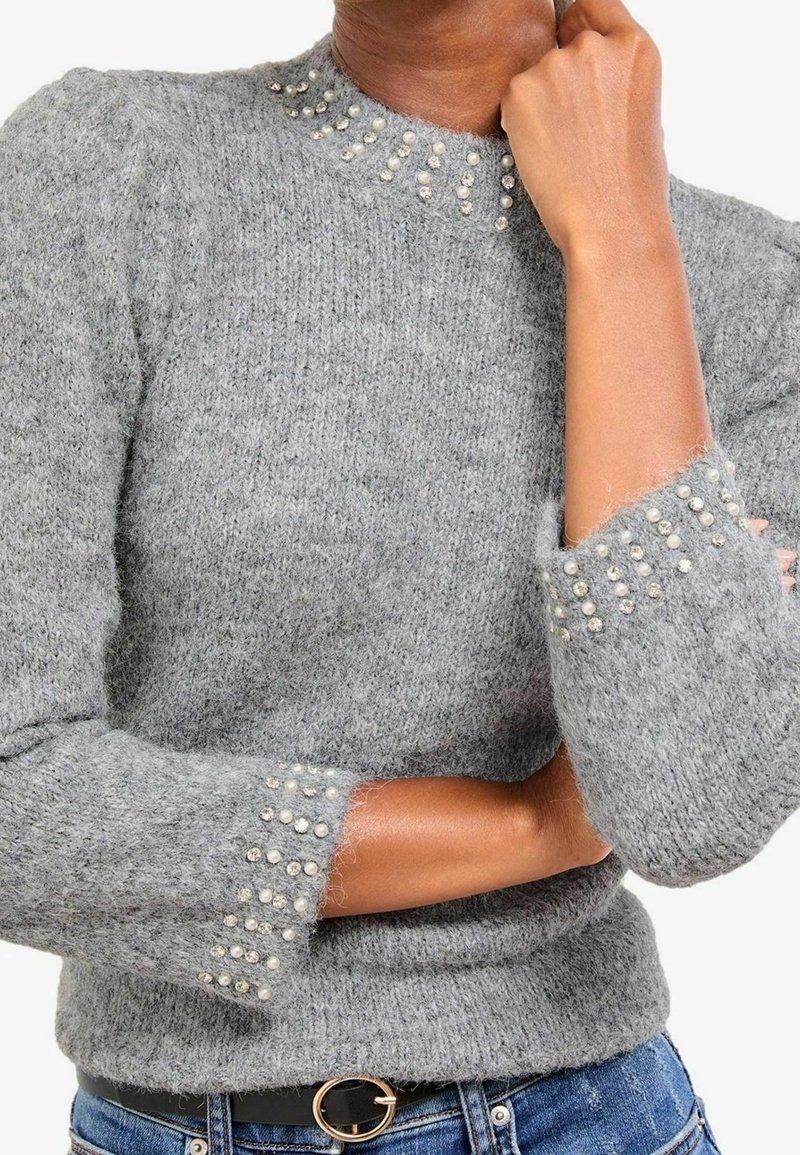 Personne portant un pull gris duveteux avec des ornements en perles et strass au col et aux poignets, associé à un jean bleu et une ceinture noire.