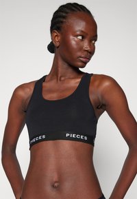 Soutien-gorge de sport noir en tissu extensible, avec décolleté en scoop et large bande élastique portant le logo "PIECES" en blanc.