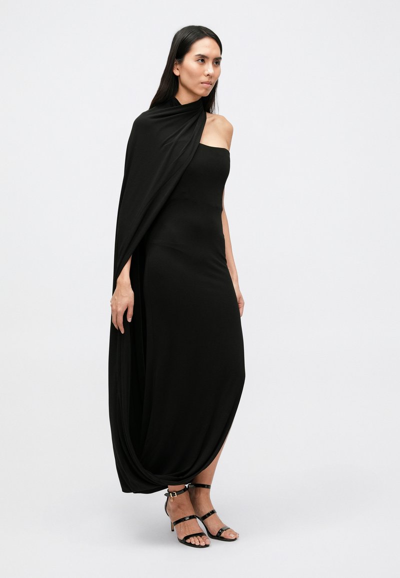 Black, ankle-length dress with a fitted bodice and flowing drape extending from one shoulder. Includes strappy black sandals.