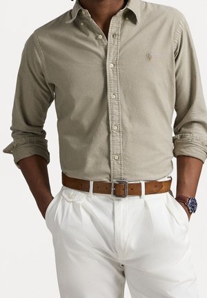 Homme portant une chemise beige à manches retroussées, un pantalon blanc plissé, une ceinture en cuir marron et une montre-bracelet avec un cadran bleu.