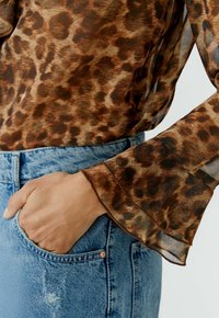 Bruine blouse met luipaardprint, gemaakt van doorschijnende stof, met uitlopende mouwen en gerafelde randjes, gecombineerd met lichtblauwe denim high-waisted jeans.