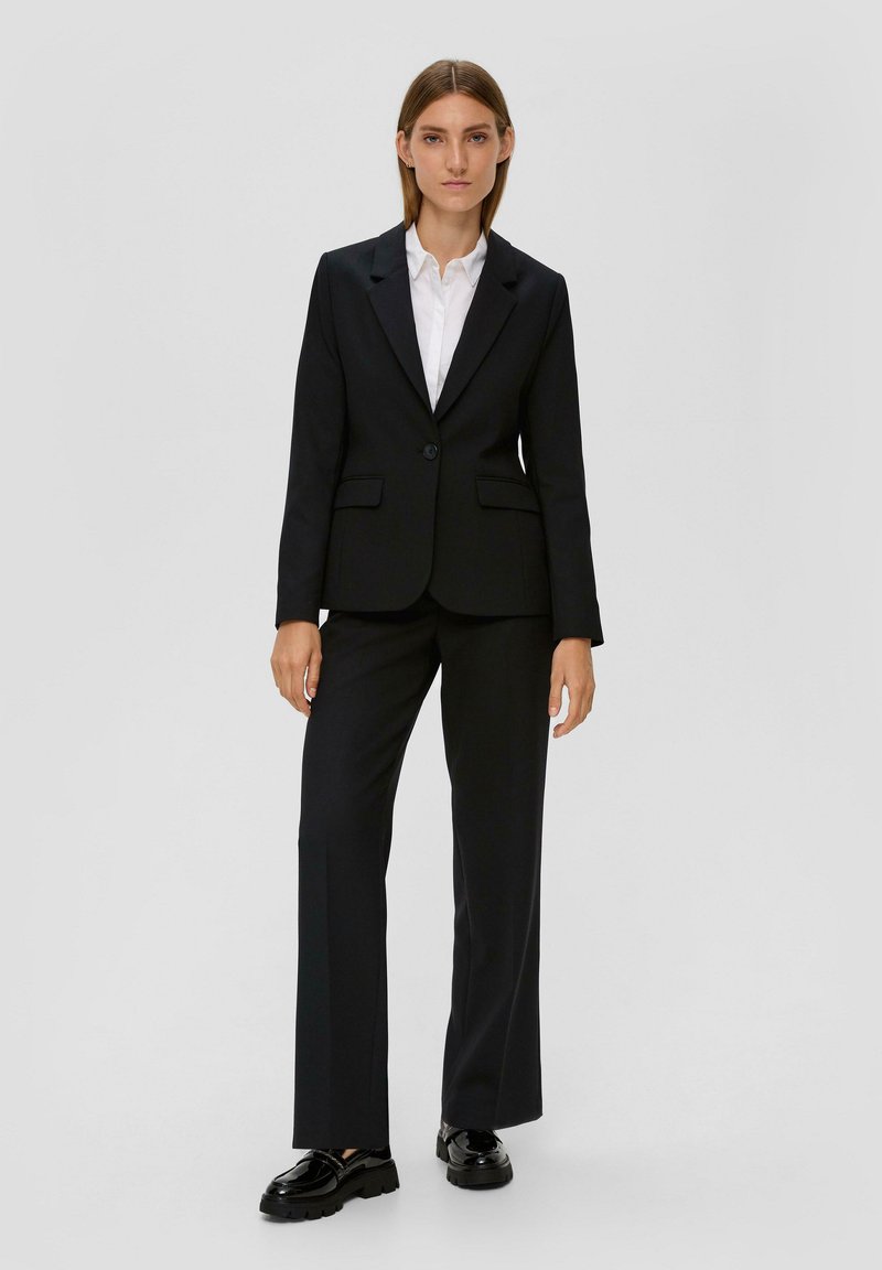 Schwarzer Anzug mit einreihigem Blazer und Revers, dazu eine passende Hose. Ergänzt wird das Outfit durch ein weißes Hemd und glänzende schwarze Schuhe.