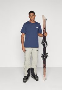 Navyblauwe T-shirt, grijze skibroek met zwarte accenten, houdt ski's vast met bindingen. Gladde stof, comfortabele pasvorm, functioneel ontwerp.