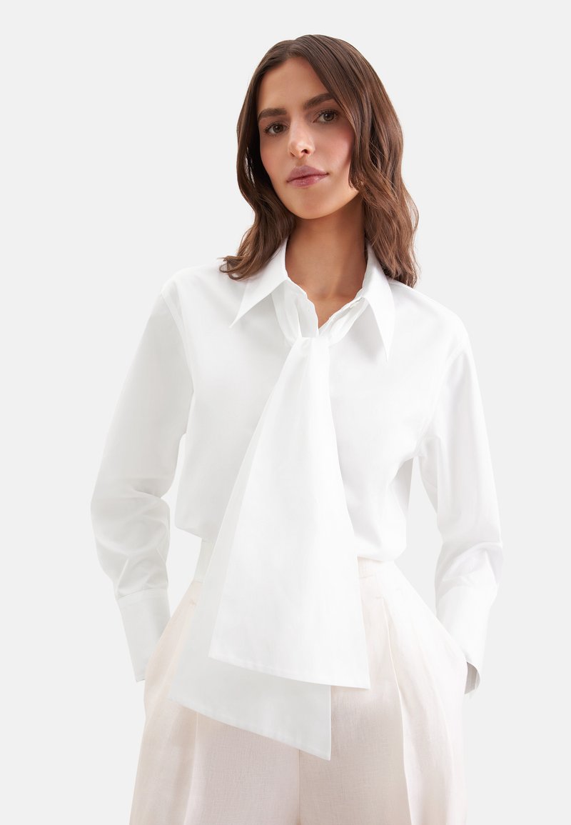 Femme portant un chemisier blanc à manches longues avec une cravate oversize, associée à un pantalon beige clair taille haute, debout les mains dans les poches.