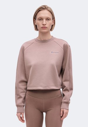 Donna con capelli biondi corti indossa una felpa corta color malva e leggings a vita alta abbinati, in piedi davanti a uno sfondo semplice.