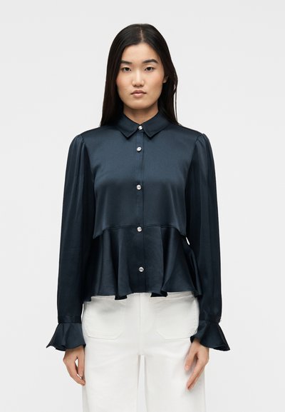 Femme portant un chemisier en satin bleu marine foncé à manches longues, avec des boutons en perle, des poignets évasés et un ourlet peplum, associé à un pantalon blanc.