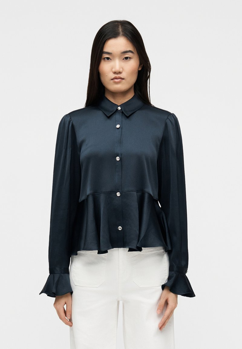 Γυναίκα που φοράει μπλούζα από σατ έν σε σκούρο ναυτικό μπλε με μακριά μανίκια, κουμπιά με πέρλες, φουσκωτά μανσέτες και βολάν στο τελείωμα, σε συνδυασμό με λευκό παντελόνι.