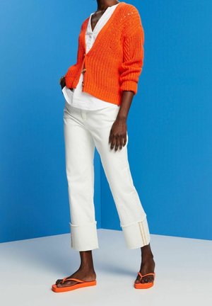 Cardigan tricoté orange avec un col en V et des boutons, porté par-dessus une chemise blanche. Associé à un jean blanc court et des tongs orange.