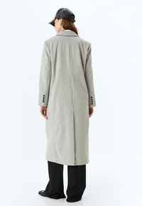 Grijze wollen jas met een lange silhouet, grote revers, knoopmanchetten en een centrale achterkantventilatie. Gecombineerd met zwarte broek met wijde pijpen.
