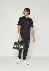 Μαύρο βαμβακερό μπλουζάκι με λευκό λογότυπο, μαύρο παντελόνι φόρμας με λευκές λωρίδες στο πλάι και μαύρη σακούλα Adidas, όλα φορεμένα με λευκά παπούτσια.