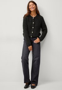 Schwarze, flauschige Strickjacke mit rundem Ausschnitt und fünf großen cremefarbenen Knöpfen, kombiniert mit dunklen weit geschnittenen Jeans und schwarzen Schuhen. Insgesamt glatte Textur.