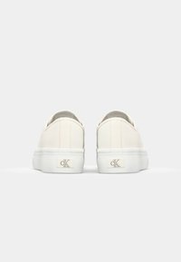 Scarpe slip-on bianche in tela con una texture liscia, dotate di una spessa suola bianca e un discreto dettaglio del logo sul tallone. Vista posteriore mostrata.