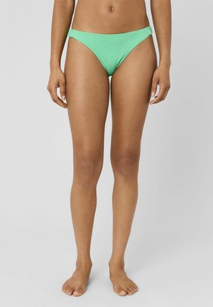Femme portant un bas de bikini texturé vert menthe, debout pieds nus devant un fond gris uni.