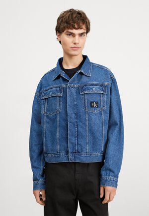 Giacca di jeans tagliata in blu medio, con due tasche sul petto, un'etichetta del logo nera e cuciture a contrasto. Maniche lunghe con polsini.