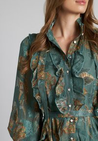 Blusa floral en tono verde azulado con cuello con volantes y detalles, que cuenta con cierres de botón y una tela texturizada con acentos de bordado en oro y verde.
