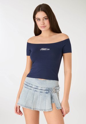 Maglietta blu navy scollata con la scritta "Tennis Club", abbinata a una gonna plissettata in denim azzurro chiaro con bottoni decorativi e design a strati.