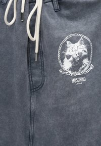 Γκρι παντελόνι από ύφασμα με λευκά κορδόνια και λευκή εκτυπωμένη σχεδίαση με μια γάτα που φορά γυαλιά και παπιγιόν μέσα σε πλαίσιο σχοινιού, με την επιγραφή Moschino Jeans.