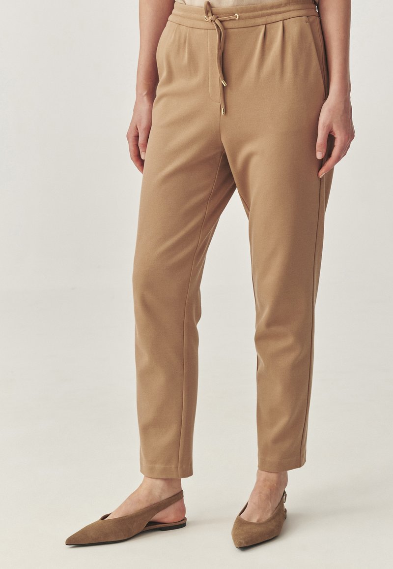 Pantaloni taper cu mai multe nuanțe de maro, cu talie elastică, șnur interior și buzunare laterale; asortați cu sandale din piele maro cu spatele descoperit. Textura materialului este moale.
