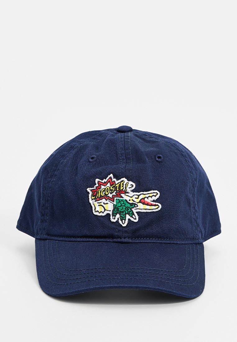 Gorra de béisbol azul marino con el logotipo de Lacoste bordado en estilo cómic, que presenta un cocodrilo verde y un estallido de texto en rojo y amarillo.